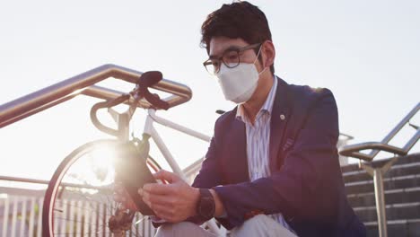 Asiatischer-Mann-Mit-Gesichtsmaske-Und-Digitalem-Tablet,-Während-Er-Auf-Der-Treppe-Im-Firmenpark-Sitzt