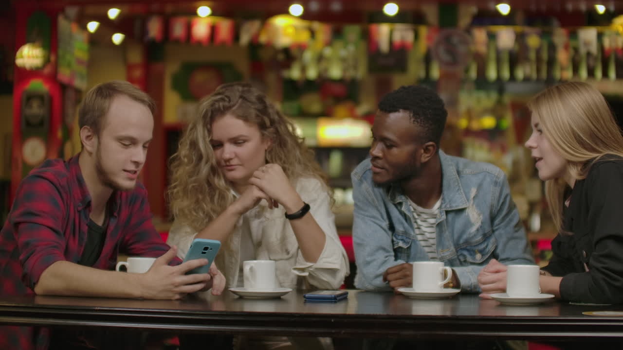 Ritratto di giovani amici allegri che guardano lo smartphone mentre sono seduti in un caffè. Persone di etnia mista sedute a un tavolo in un ristorante che usano il telefono cellulare.