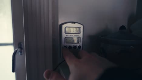 hand pushing buttons of a wall-mounted digital thermometer measuring indoor temperature at home