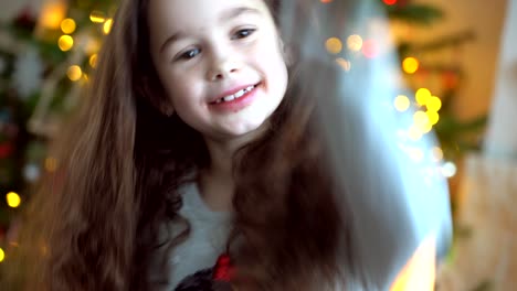 portrait of happy little girl sitting near christmas tree on new year's eve in decorated room. new year and holidays with kids concept. enjoying festive holiday celebration.