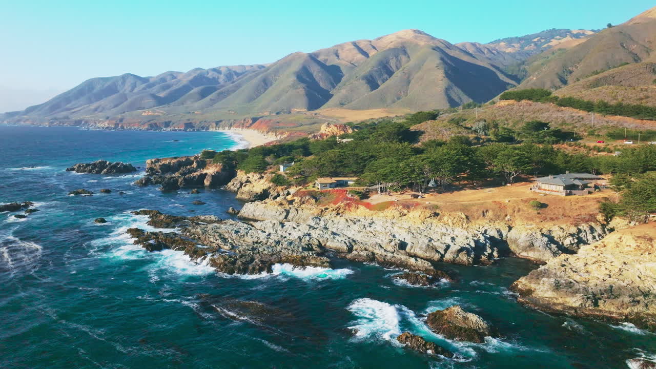 Houses among green trees growing at the rocky coast of California. Majestic bare mountains at the backdrop of blue clear sky.