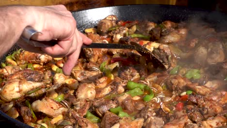 a chef stirs a meaty spanish paella as it is being cooked in a large saucer pan, close up, slow motion view shot from the right side of the cook