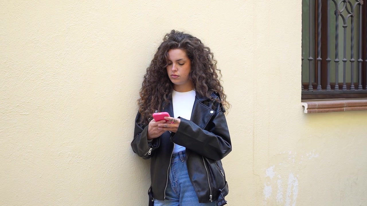 Una giovane donna elegante con i capelli ricci sta usando il suo smartphone vicino a un muro giallo brillante.
