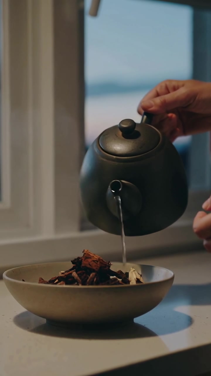 Wideo pionowe: Przechylający się czajnik, ręce nalewające wodę do miski z ziołami na herbatę na blacie kuchennym, szlafrok