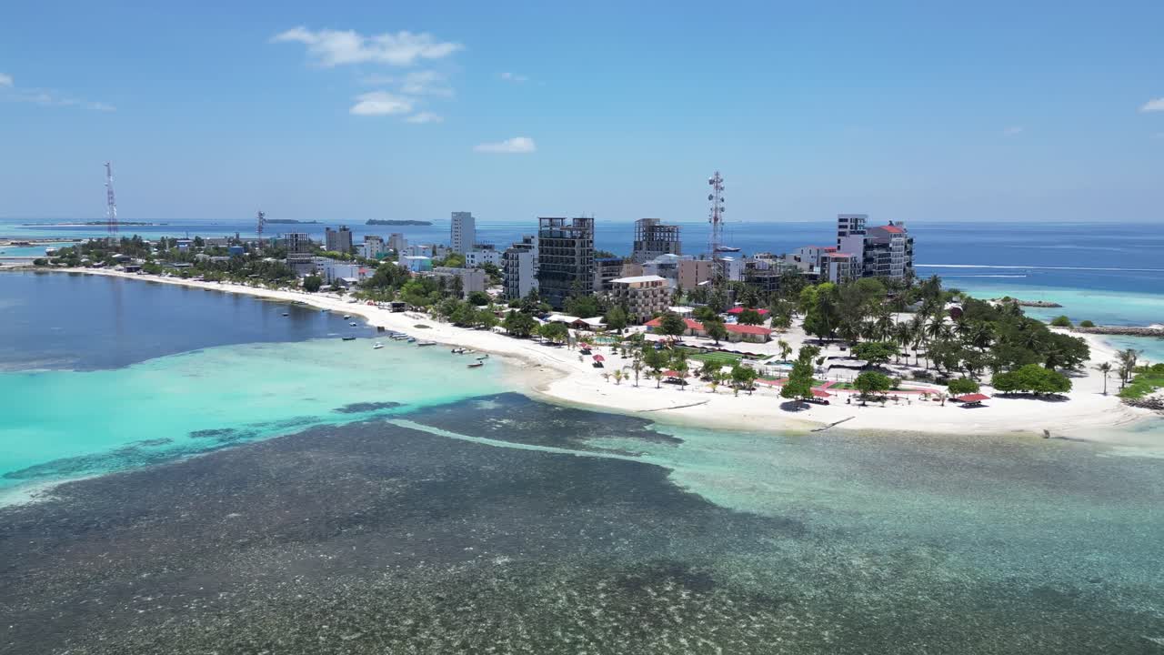 Aerial view of tropical coastal town with white sandy beaches and clear turquoise water, showing urban buildings along the shoreline. Maafushi Maldives.