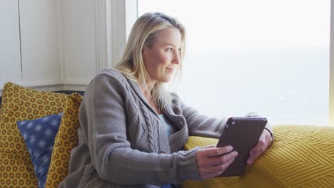 Glückliche-Kaukasische-Reife-Frau,-Die-Durch-Das-Fenster-Schaut-Und-Ein-Tablet-Benutzt