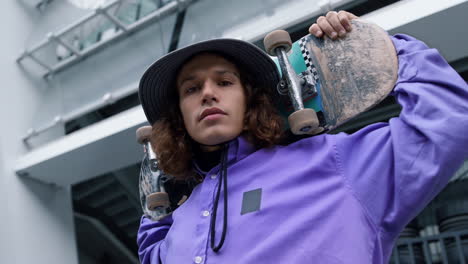 Serious-skater-holding-skate-on-shoulders.-Focused-guy-looking-camera-outdoor.