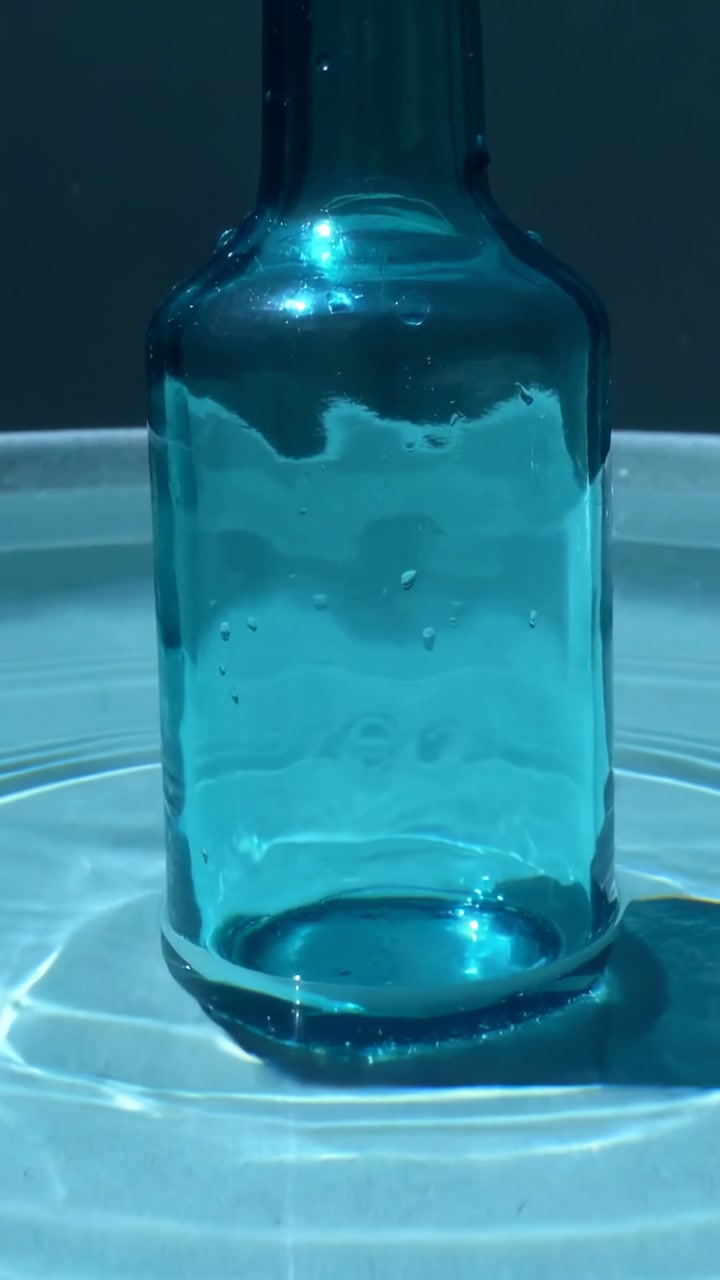 Vertical video: Rippling rings causing teal bottle casting glow in basin with droplets shifting