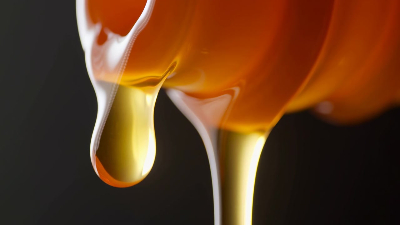 Scena macro di liquido ambrato viscoso che forma goccioline lucide accanto a un flusso più denso, con una gocciolina che si arrotonda liberamente mentre la colonna adiacente si assottiglia sotto una calda luce da studio.