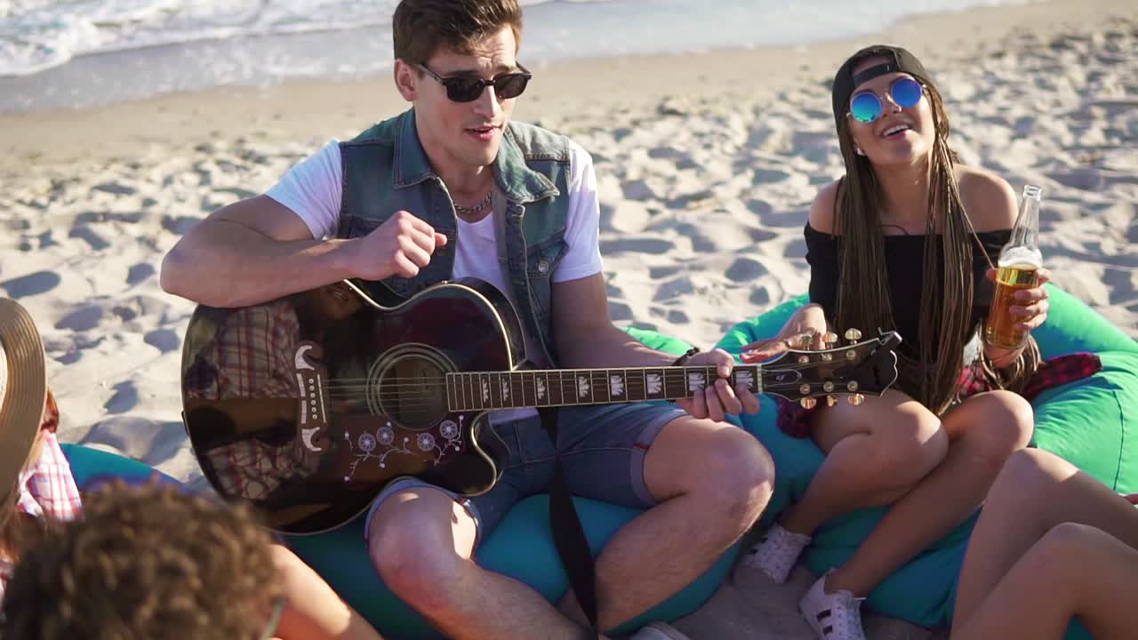 joven tocando la guitarra entre un grupo de amigos sentados en sillas fáciles en la playa y cantando en una noche de verano. disparo en cámara lenta