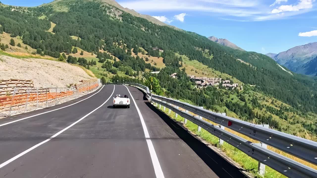 A car travels on a scenic mountain highway, surrounded by lush greenery and clear blue skies.