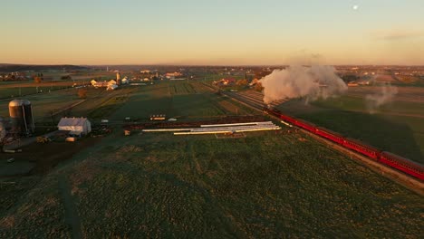 drone view from above and behind of a steam engine approaching blowing lots of smoke at sunrise traveling thru the farmlands on fall morning