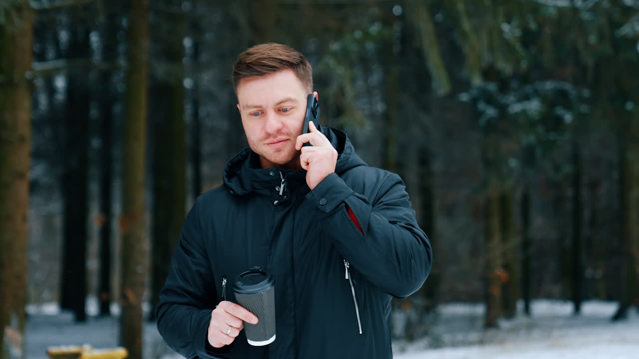Dark-haired male in black jacket walks by the winter nature. Man speaks on the phone and drinks coffee on the go.
