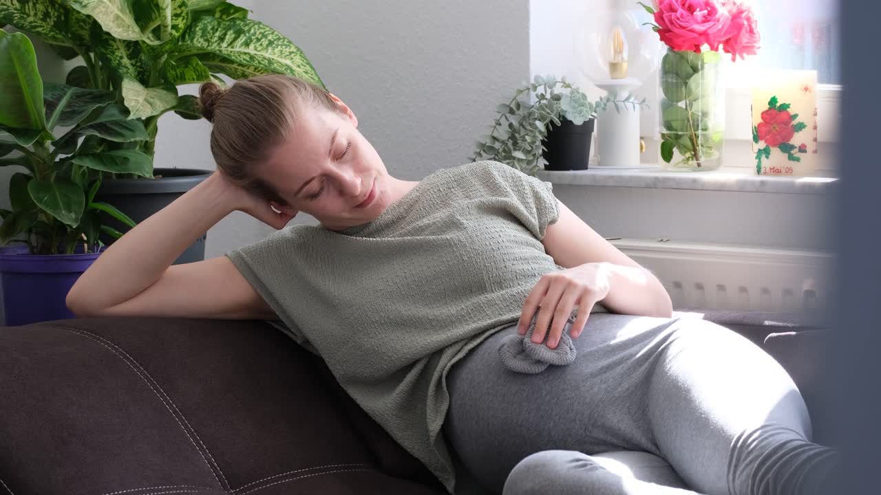 Pregnant white woman sitting on couch, playing with grey gloves on her belly smiling slowmotion
