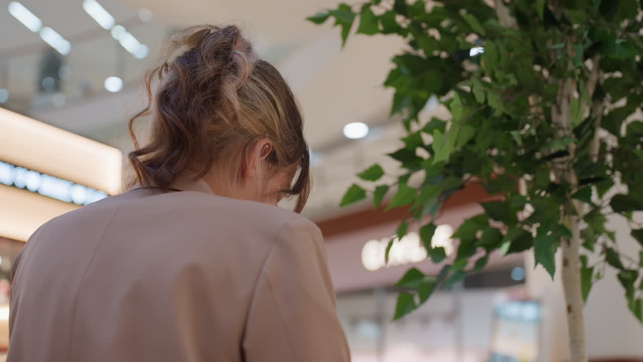 쇼핑객들 사이에서 생각에 잠긴 젊은 여성, 실내 녹지 근처에서 메모를 읽는 백인 여성, 녹지 및 조명이 있는 활기찬 쇼핑몰에서 메모를 신중하게 검토하는 백인 여성