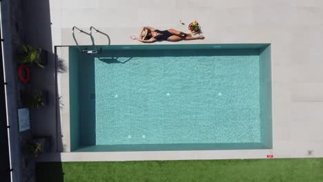 Dark-haired-slim-woman-lying-at-the-edge-of-a-beautiful-outdoor-pool---she-wears-a-dark-swimming-costume-and-enjoys-the-sun---drone-films-her-from-a-bird's-eye-view