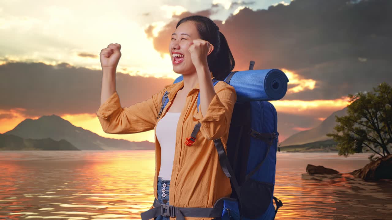 vista lateral de una excursionista asiática con una mochila de montañismo gritando la meta celebrando el éxito mientras está de pie en un lago durante el atardecer