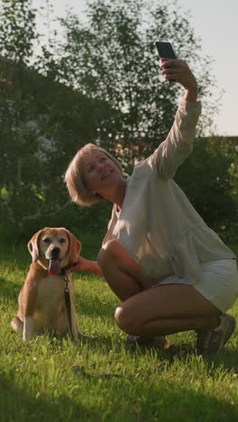 woman taking selfie with her happy dog outdoors in lush green garden under sunny weather, with building and trees in background, dog is sitting, panting joyfully, as owner holds phone smiling