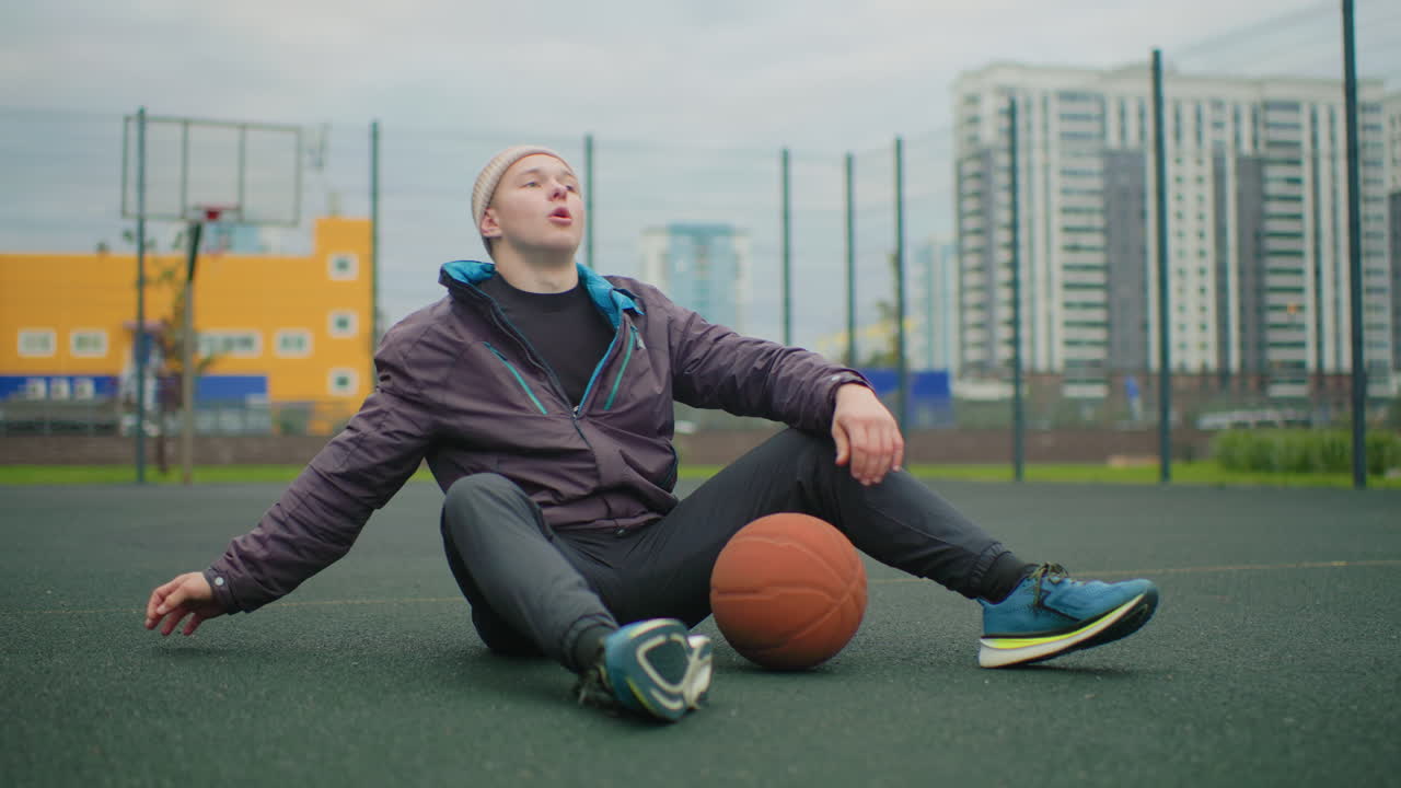 young athlete sitting on basketball court, holding orange ball, city apartment towers backdrop, overcast sky, purple jacket and headband, relaxed posture with hand behind, sneakers on asphalt, quiet