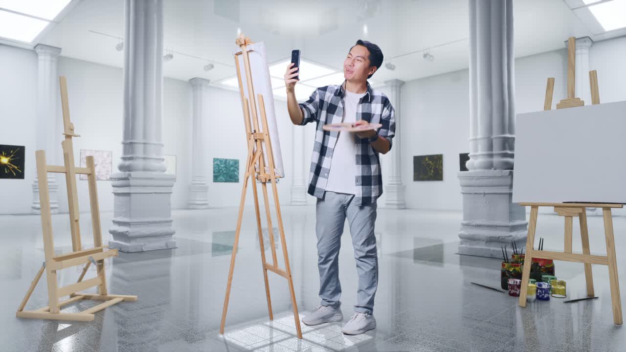 갤러리에서 오일 페인트와 브러쉬로 캔버스에 그림을 그리는 동안 스마트폰에서 비디오 통화를 하는 아시아 남성 예술가의 몸이 가득 차 있습니다.