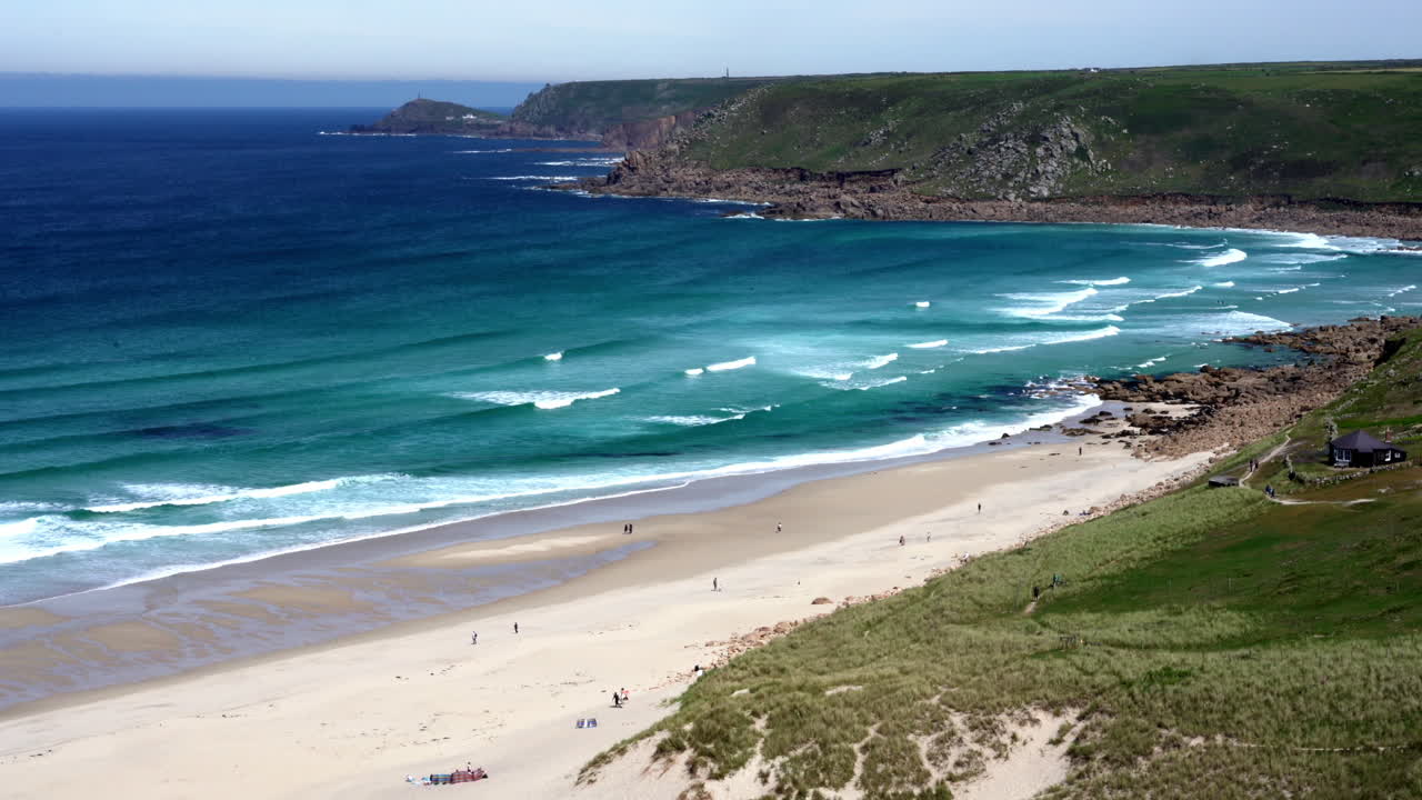 Meren aallot leveällä hiekkarannalla Sennen Coven poukamassa Cornwallissa, surffaajien ja turistien kävellessä rannalla