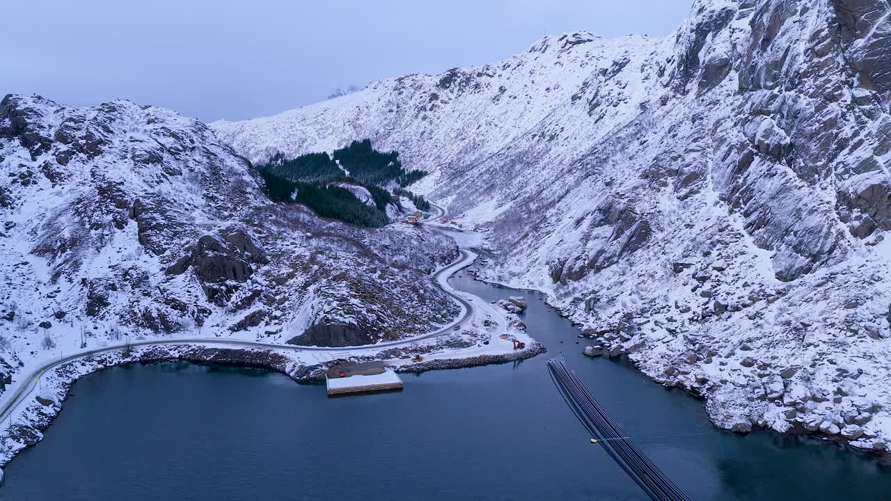 Eine atemberaubende Luftaufnahme von schneebedeckten Gipfeln, einer gewundenen Küstenstraße und tiefblauem Fjordwasser auf den Lofoten, Norwegen. Ideal für Reise- und Naturdokumentationen.
