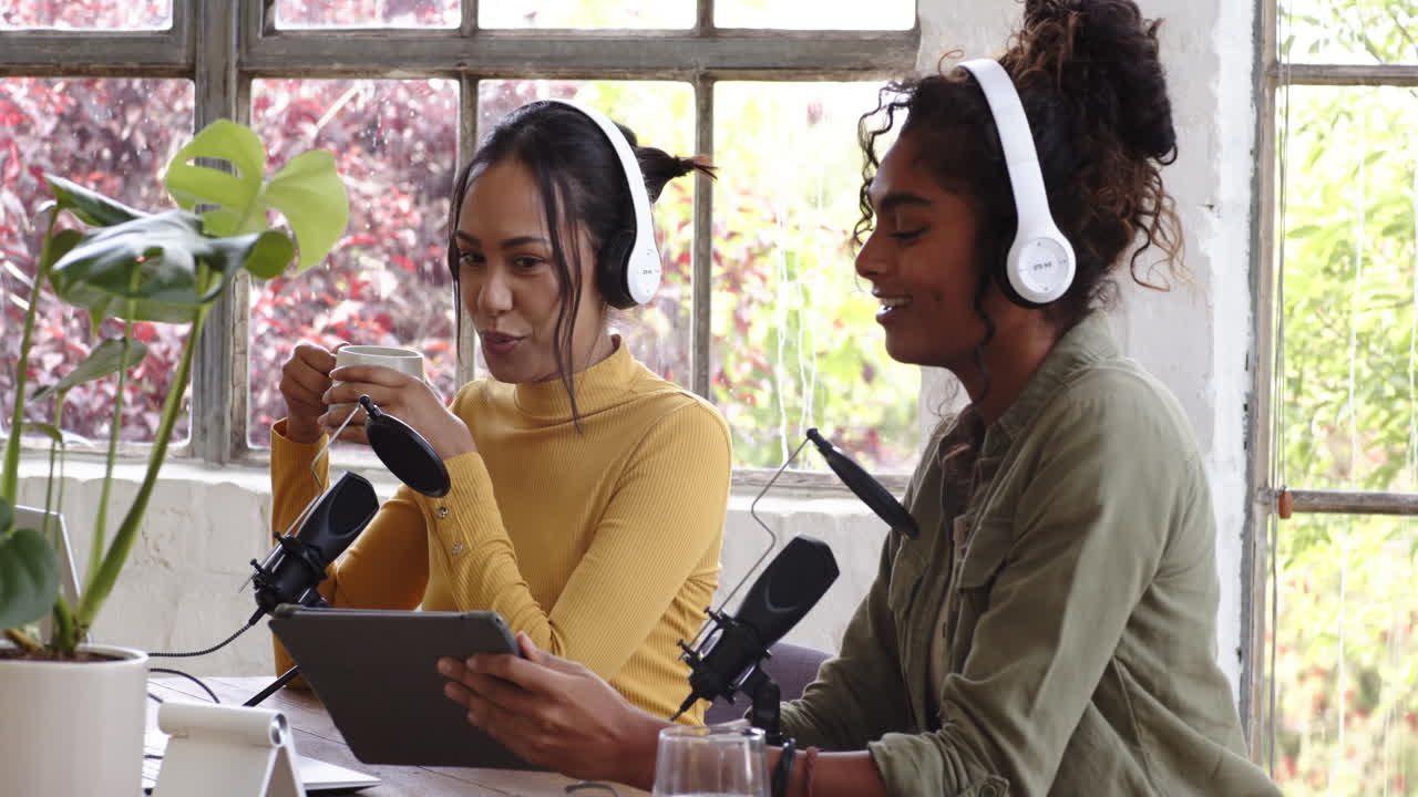 Two multiracial female colleagues podcasting at home, wearing headphones and using microphones