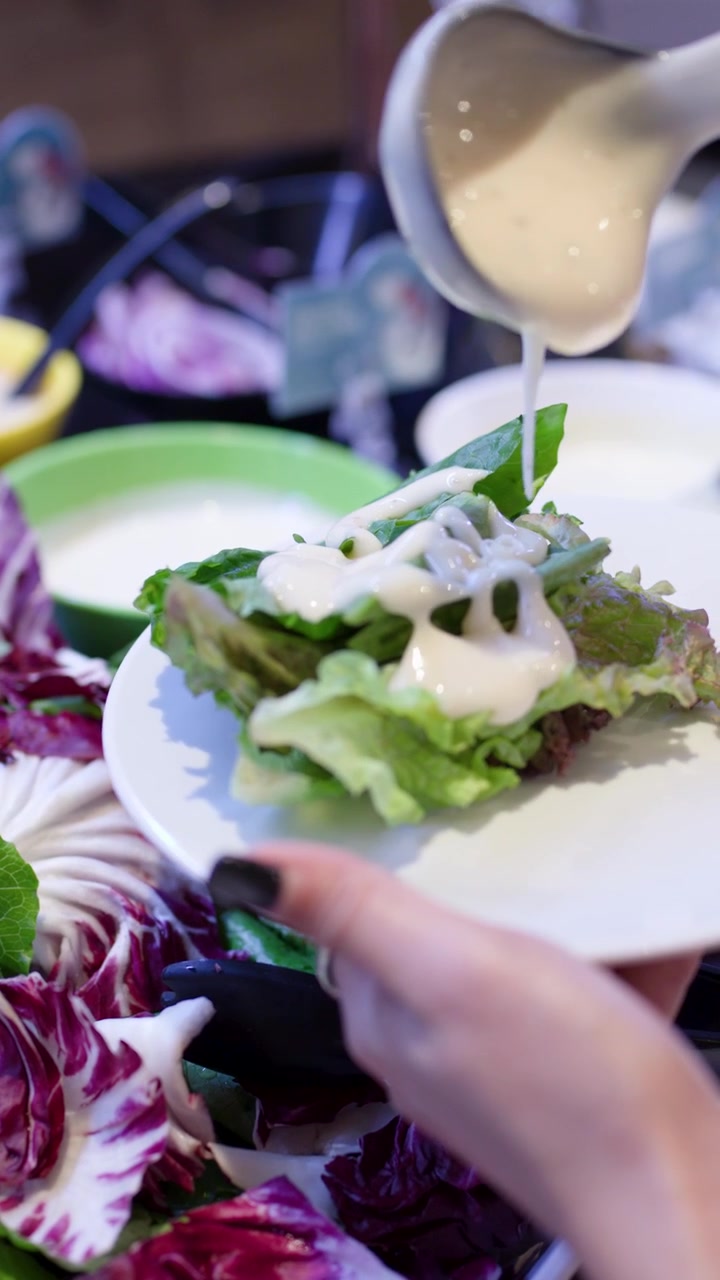 Nærbillede af en hånd, der øser cremet dressing over grøn salat ved en farverig buffetstation.