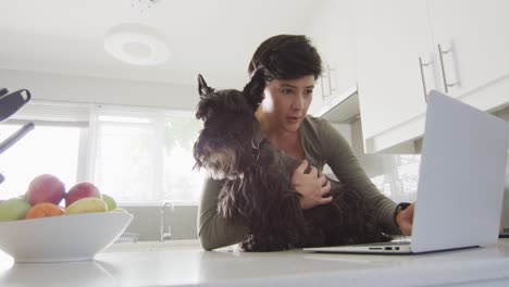 Mujer-Caucásica-Sosteniendo-A-Su-Perro-Y-Usando-Una-Computadora-Portátil-En-Casa