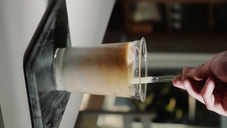 video vertical home barista revolviendo café y leche para crear un café con leche helado en un vaso de vidrio con pajita de vidrio en cámara lenta de cerca