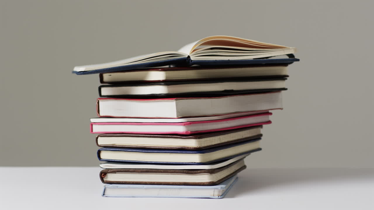 A stack of various hardcover and paperback books sits against a neutral background, with copy space