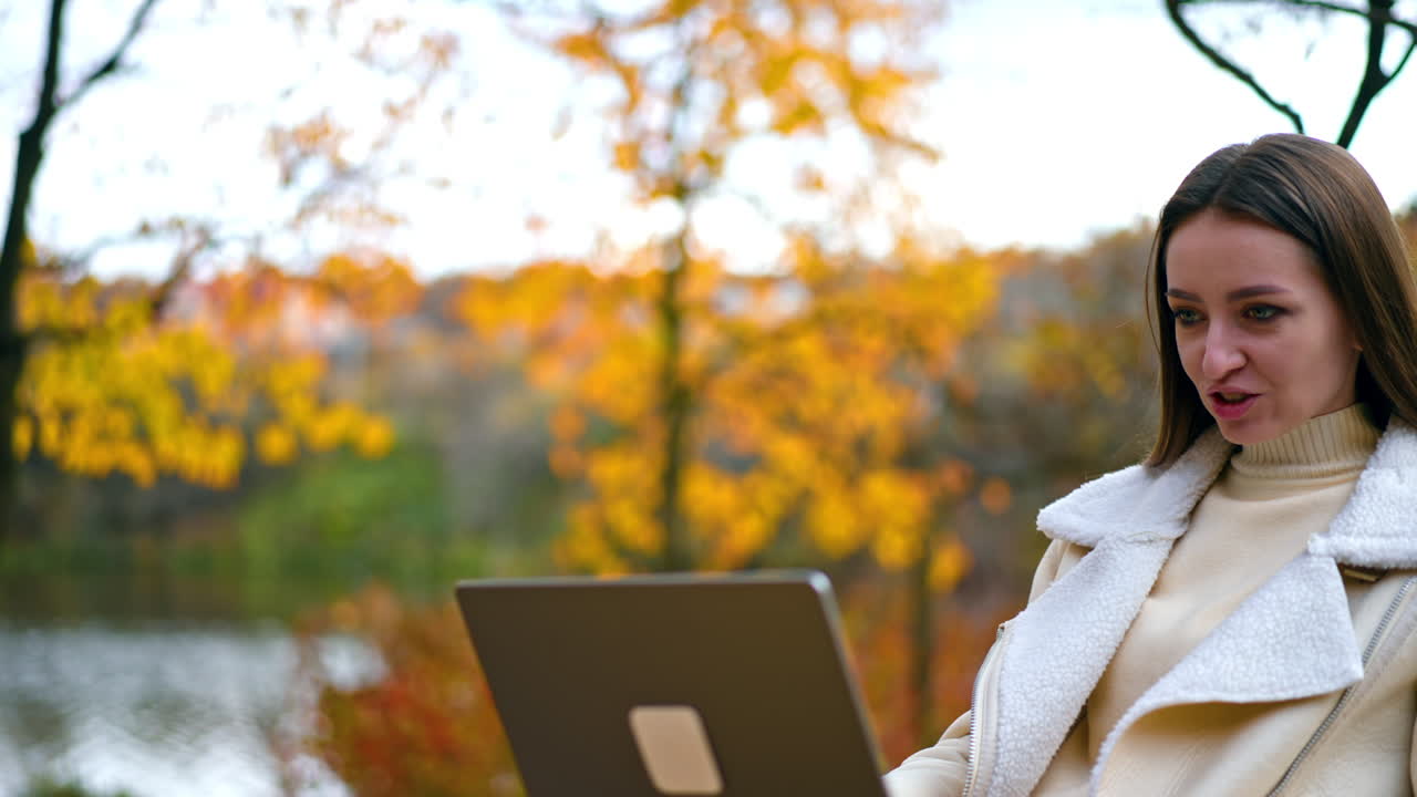 Happy smiling woman looking at the screen of her laptop sitting outdoors. Brunette lady using computer resting in the autumn park.
