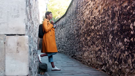 Young-woman-wearing-a-yellow-pea-coat-and-blue-jeans-leaning-on-stone-wall-in-an-alleyway-talking-on-her-smartphone-using-earphones,-low-angle,-full-length