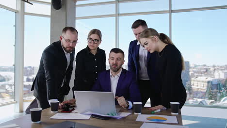 experienced businessman sits at desk with laptop and his staff around him. leader shows something at computer to his team. panoramic windows with cityscape at backdrop.