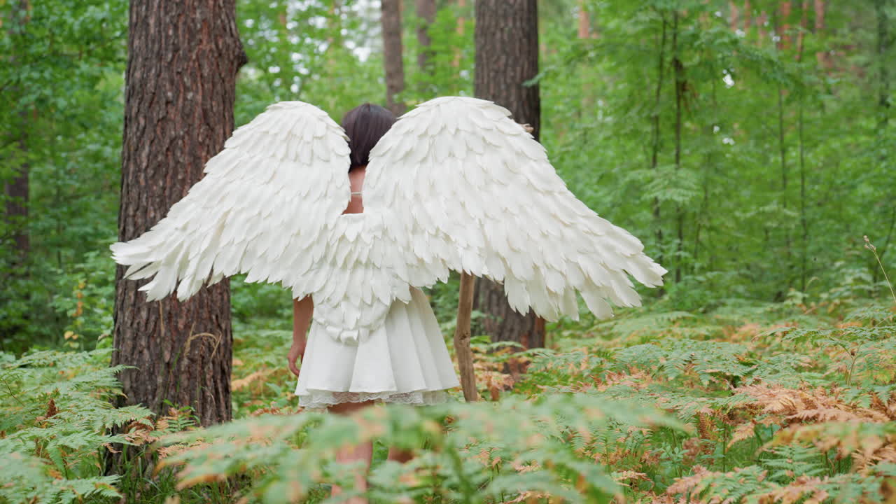 Rear view of young woman dressed in white angel costume walking through lush woodland, holding stick and gently touching green plants, surrounded by tall trees and soft light
