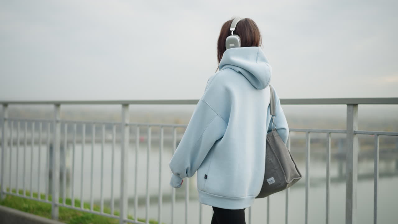 Side view of beautiful girl in casual hoodie walking with bag near iron rail, blurred view of bridge and river in background, creating a calm, urban atmosphere perfect for lifestyle content