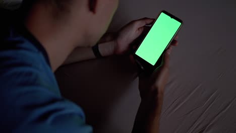hombre en la oscuridad en la cama sostiene y examina el teléfono inteligente con pantalla táctil verde