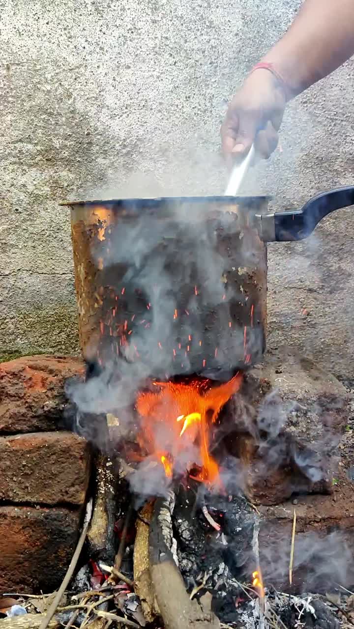 In an open space in Maharashtra, India, food is being prepared on a traditional chulha. Wood crackles in the fire beneath a metal cooker, as smoke swirls gently in slow motion, evoking rustic charm