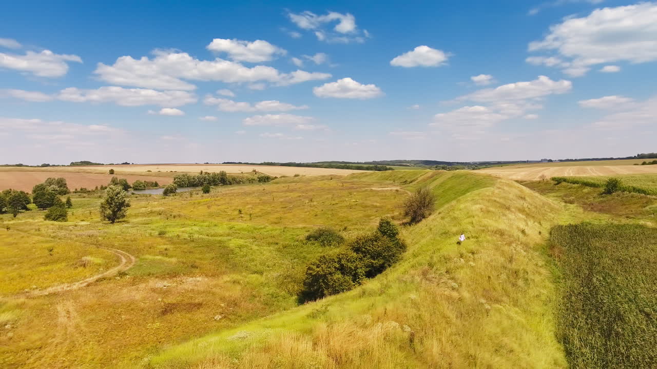 Woman in white dress walks along the hill. Sunny meadow bordering with farmlands. Bright blue sky with white clouds at backdrop.