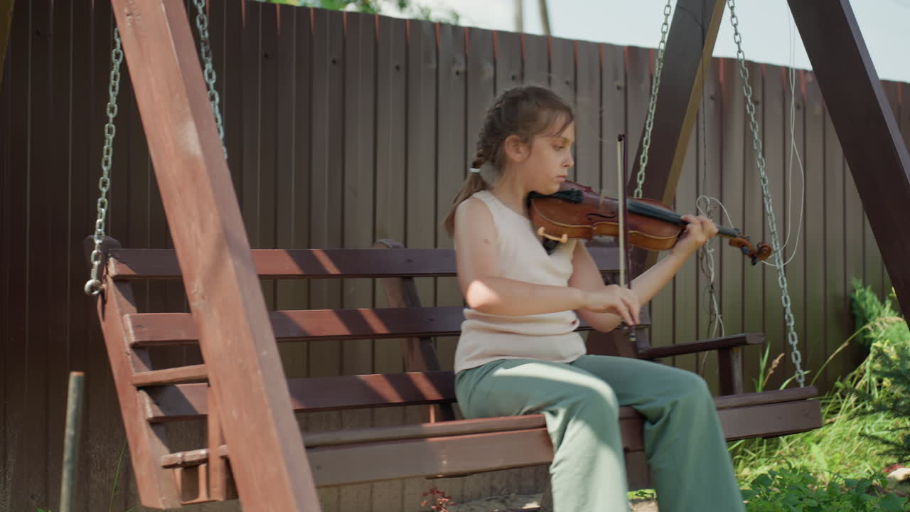 Young Girl Outdoors With Violin, Girl With Bow In Sunlit Garden Playing Violin On Swing, Casual Summergear Girl Attentively Practicing Violin Outdoors Amidst Lush Greenery And Fencing