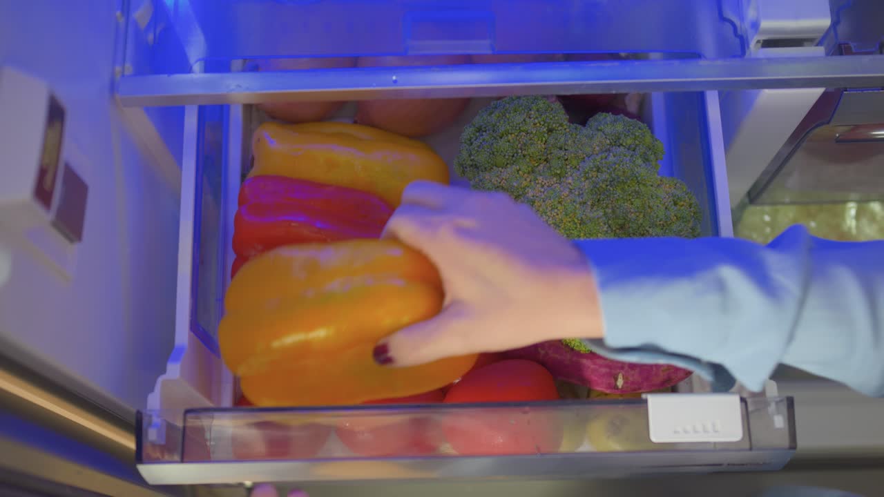Eine Person schaut in einen Kühlschrank und holt Gemüse heraus. Das Gemüse umfasst Brokkoli, Paprika und Karotten.