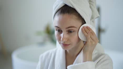 Portrait-of-beautiful-girl-in-bathrobe-and-with-towel-on-her-head-cleaning-her-face-with-sponge