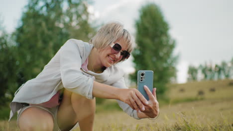 dama de mediana edad con gafas de sol agachada mientras toma una foto con una cálida sonrisa en un vasto campo abierto con árboles en el fondo borroso, la luz natural del sol destaca su expresión alegre