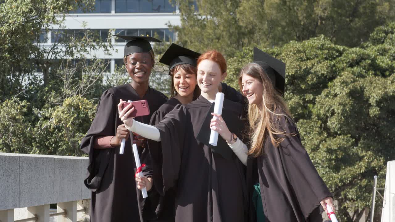 Diversas mujeres graduadas con birretes y togas haciéndose un selfi con sus diplomas y un teléfono rosa en una terraza