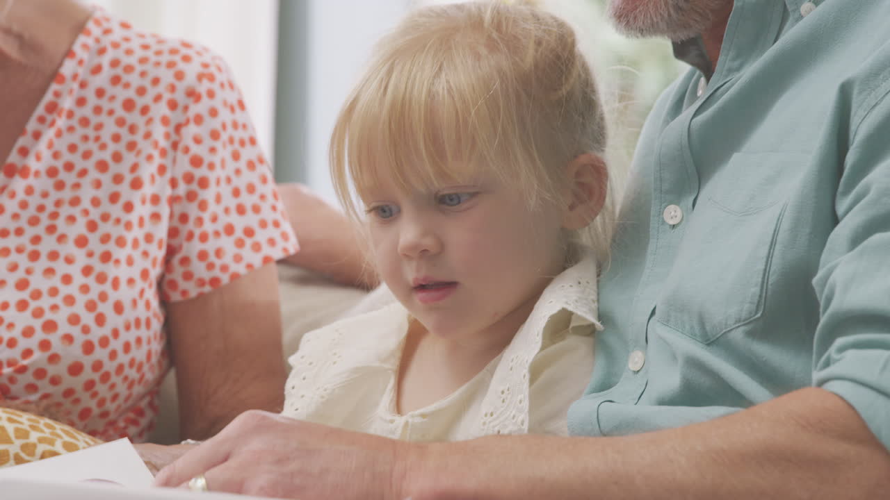 집 에서 손녀 와 함께 소파 에 앉아 함께 책 을 읽고 있는 할아버지 와 할머니 의 가까운 모습