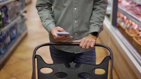 Man-using-mobile-phone-while-shopping-in-supermarket,-trolley-mall-grocery-shot-store