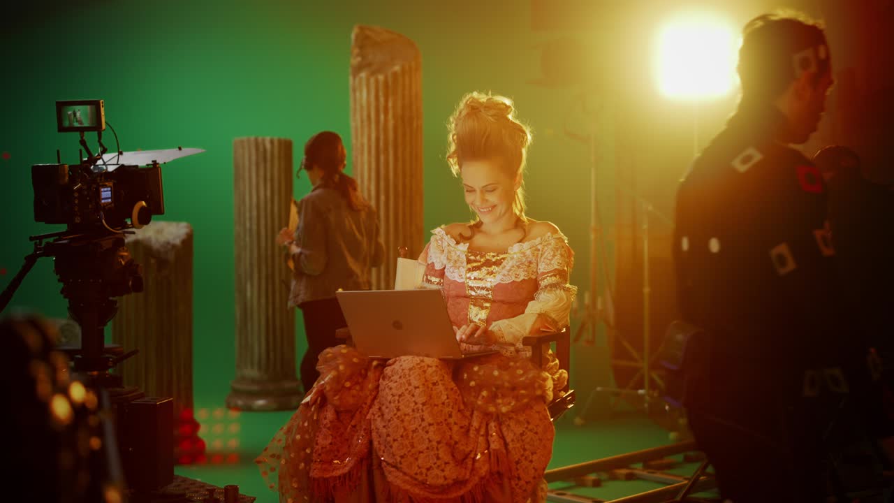 en el set de películas de drama de época: hermosa actriz sonriente con un vestido renacentista, sentada en una silla usando una computadora portátil con pantalla verde en el fondo. historia de rodaje de estudio blockbuster