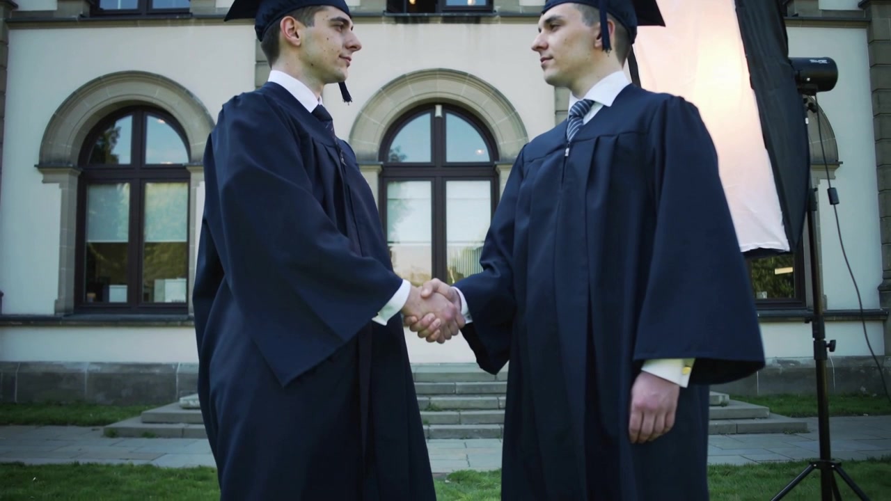 Extending hands two grads shaking hands on campus, in gowns and caps for graduation, softbox