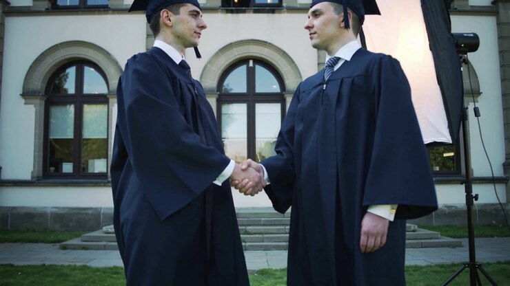 Due neolaureati che si stringono la mano nel campus, in toga e tocco per la laurea, con softbox.