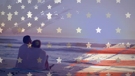 Animation-of-american-flag-over-happy-diverse-couple-wrapped-in-rug-embracing-sat-on-sunset-beach